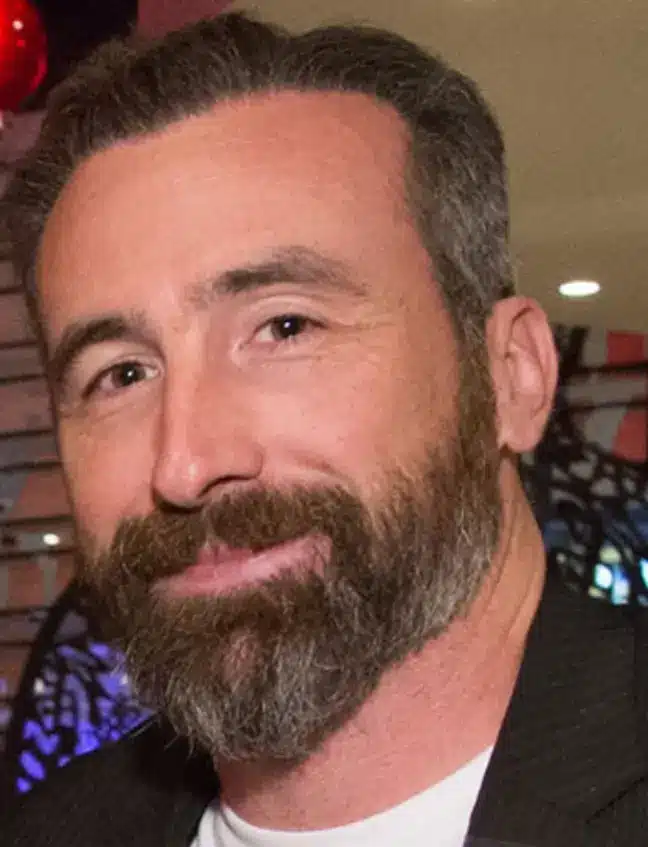 Man with beard smiling at camera in indoor setting with dark background and lighting.