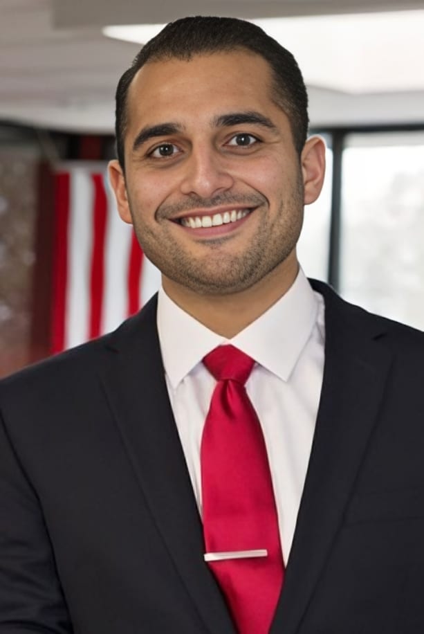 Professional man in dark suit and red tie smiling in modern office setting with American flag